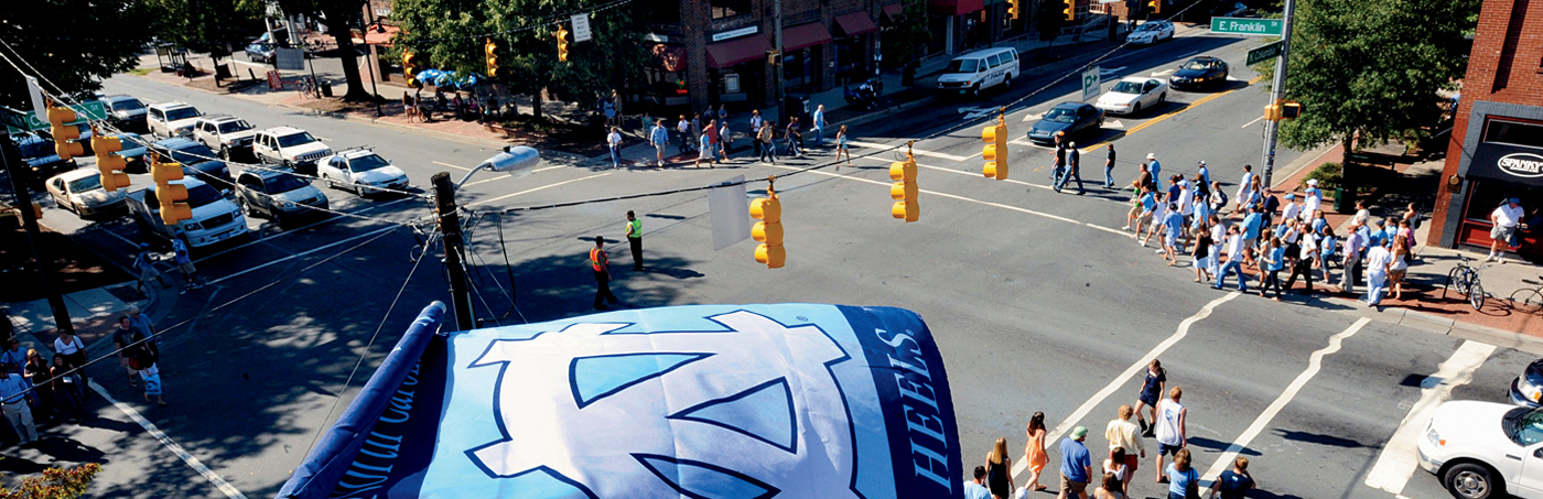 Students and fans cross an intersection in Chapel Hill, NC under a Tar Heels flag.