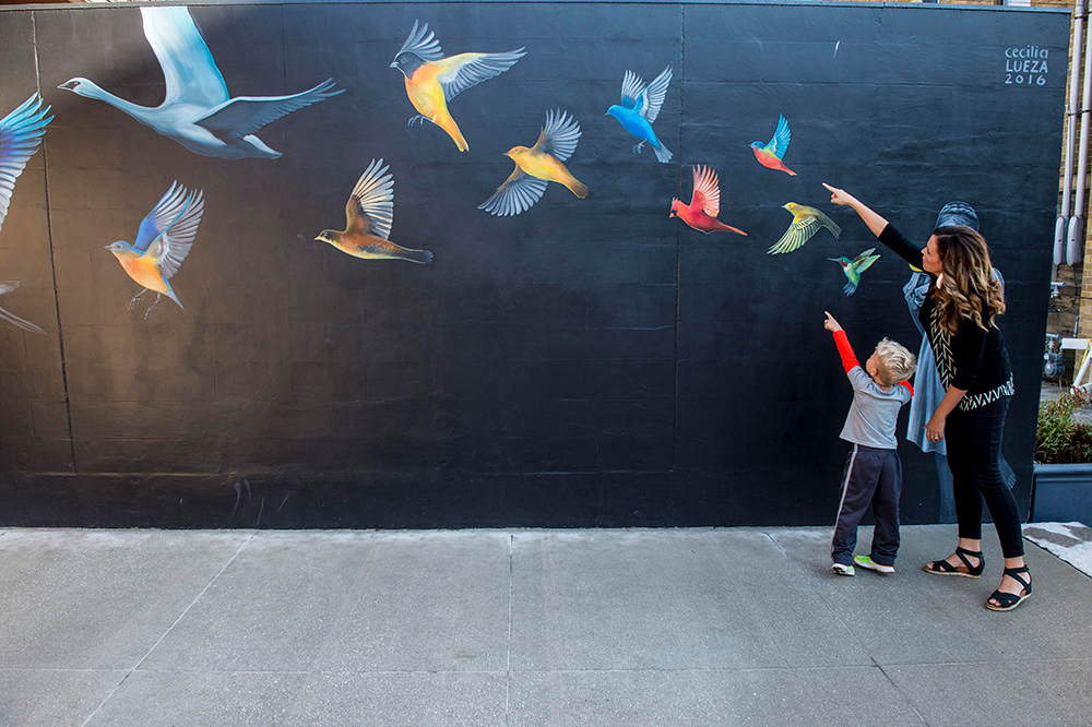 ImaginArt in the Alleys Angie Glick is the woman in the red top and shopping bag underneath the archway entrance. She was later joined by Brooke Prouty (blue sweater) and Emily Ruff (black, gold and black striped top). Pouty and Ruff were photographed examining a ceramic pot in “Be Inspired” a home decor shop owned by Jill Deklotz. The three models then moved to an adjacent store “Di Moda” where they tried on hats and inspected goods with assistance from owner Tammy Cannavo (dark streaked hair standing behind the display as Glick tries on a bracelet). The last shop visited was Joy Boutique. Prouty was assisted with the sizing of a dress by boutique owner, Joy Purdy. The next shots of the guy in the checked shirt looking and taking photos of some of the artwork is local architect Kyle Martin of Martin Gardner Architecture in Marion. The next shots underneath the archway are of Marion resident Ben High with his daughter, Lucy. I do not have names of the people drinking on the back patio of Uptown Snug. The next people shots are of the two women and dog along downtown store fronts. The woman with the dreads and scarf is Gae Sharp-Richardson. The woman in the purple top is Sarah Presley. The dog’s name is Luna. The next shots are of a guy in a blue checked shirt and a woman in a black sweater. The man’s name is Will Brandt. The woman’s name is Katrin Elessais. The next shot is of two women and a little boy looking in the store windows. The woman in the dress and boots is Tami Mysak. The other woman is Joni Leymaster with her son, Zac. The woman with short curly hair and pink top is Cathy Petersen (owner of Wit’s End Coffee House). The guy looking in the store windows with Petersen is Dan Washburn. The tall glass sculpture that Martin is looking at is titled “Prime Commonality” by Luke Crawley and Quincy Jones. Martin was also looking at a section of “Drawing Room” by Matthew Kargot. The metal sculpture that Prouty and Ruff are pictured under is "Alley Blome" by Jake Falcom. The entrance that Glick is under is “Prairie Schooled” by Cara Briggs Farmer. The fist sculpture with the green bike (courtesy of Cranky’s Uptown Bicycles) is titled “Life is a Ride” by Chris Miller. The bird painting that Zac and his mother, Joni are photographed in front of is “Midnight Wonder” by Cecilia Lueza. The sculpture that Sharp-Richardson and Presley are sitting underneath is “Evoke” by Dale Merrill. The industrial-looking sculpture shot from above with Brandt and Elessais looking on is “Confluence” by Dan Perry.