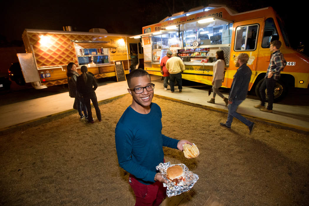 A man in a blue shirt holds a burger and fries that he got from a food truck behind him in Denton, TX.
