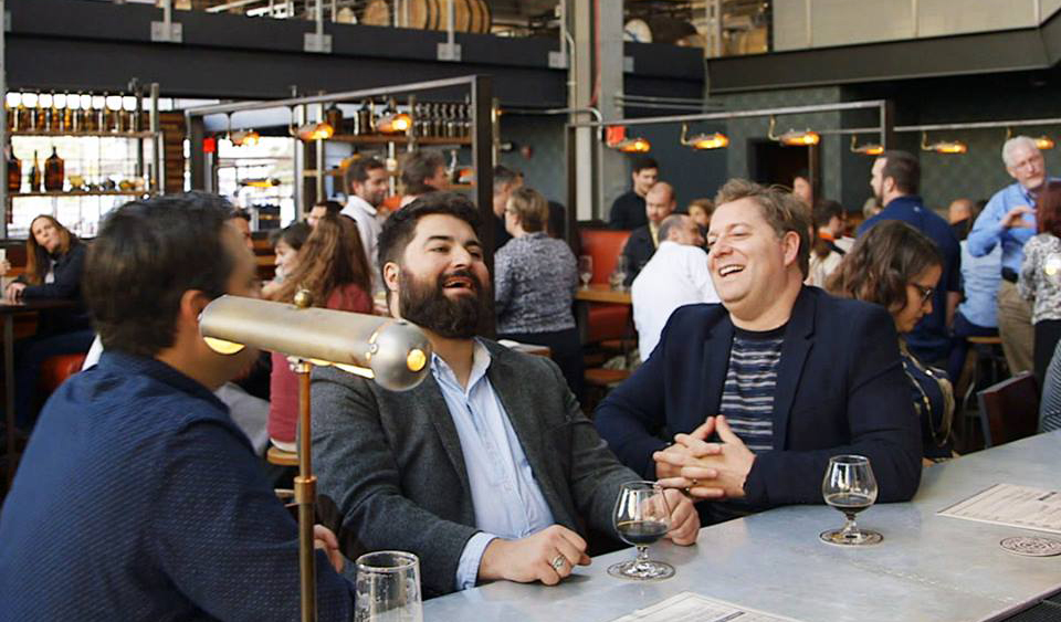 A bearded man laughs while drinking at the bar with friends at BlueJacket Brewery.
