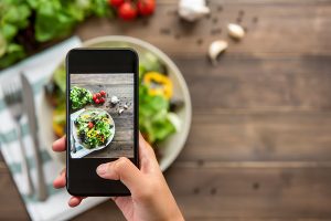 Food blogger using smartphone taking photo of beautiful mix fresh green salad on wood table to share on social media