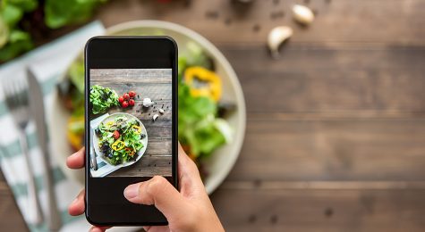 Food blogger using smartphone taking photo of beautiful mix fresh green salad on wood table to share on social media