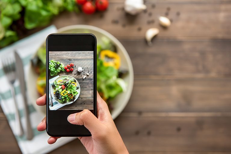 Food blogger using smartphone taking photo of beautiful mix fresh green salad on wood table to share on social media