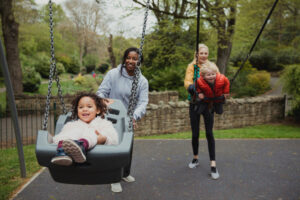 Two mothers are pushing their son and daughter on the swings in the park. Mom friends make raising children more fun.