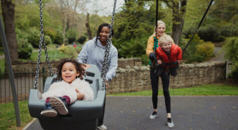 Two mothers are pushing their son and daughter on the swings in the park. Mom friends make raising children more fun.