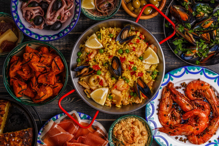 Typical spanish tapas concept. Concept include slices jamon, bowls with olives, anchovies, spicy potatoes, mashed chickpeas, shrimp, calamari, manchego with quince marmalade, pans with tortilla, paella, mussels on a wooden table.