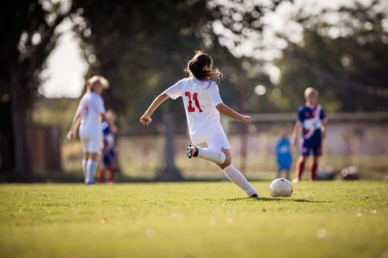 Back view of female soccer player kicking the ball during a match on a stadium. These cities support women's sports.
