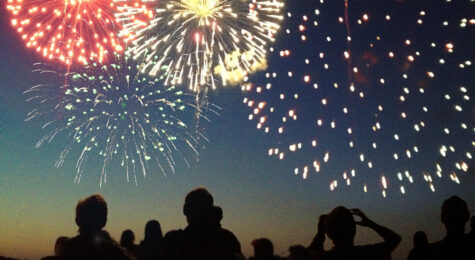 A Fourth of July fireworks display exploding in the sky over a group of people. The crowd, spectators in silhouette, celebrate the Independence Day national holiday together, looking up and watching bursts of color. The pyrotechnic performance may represent New Year’s Eve, Chinese New Year, or other traditional festival celebration events.