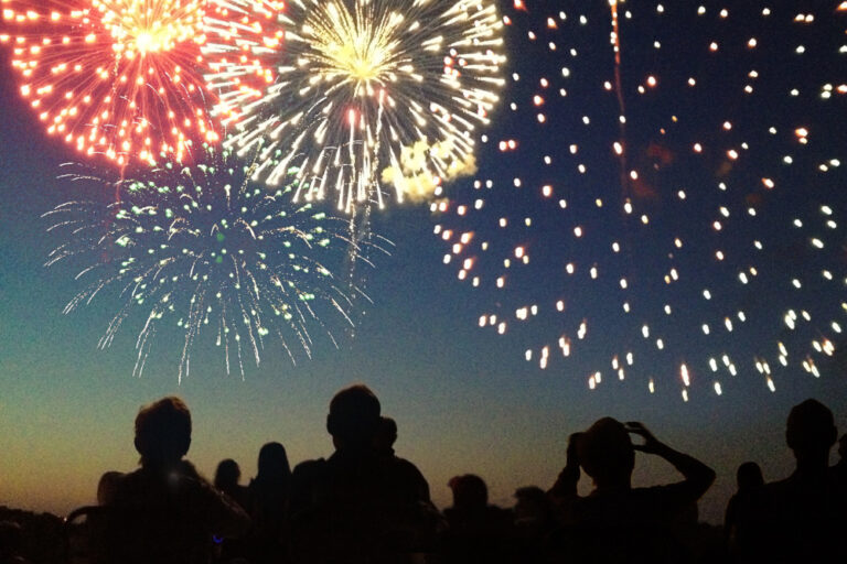 A Fourth of July fireworks display exploding in the sky over a group of people. The crowd, spectators in silhouette, celebrate the Independence Day national holiday together, looking up and watching bursts of color. The pyrotechnic performance may represent New Year’s Eve, Chinese New Year, or other traditional festival celebration events.